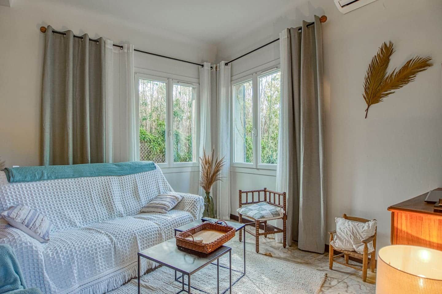Ein gemütliches Wohnzimmer mit einer Couch, einem kleinen Tisch und zwei Sesseln. Große Fenster mit Vorhängen, eine Lampe und eine Wanddekoration aus einer Feder sind zu sehen.