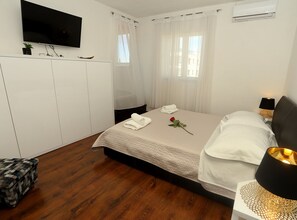 A hotel room with a bed, a TV mounted on the wall, a bedside table with a lamp, and a wardrobe. - Apartments Villa Di (Okrug)