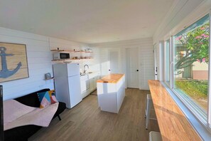 Une cuisine compacte avec des meubles blancs, un four à micro-ondes et un évier. On y trouve un plan de travail en bois et une fenêtre donnant sur la verdure à l’extérieur.