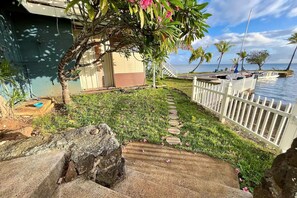 A coastal property with a pathway leading to a white picket fence, a small garden with pink flowers, and a view of the sea. - Kaneohe Bay 2BR - Oceanfront Pier & Kayaks - Wi-Fi (Kaneohe)