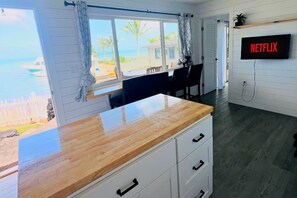 A room with a wooden counter, a white cabinet, a TV mounted on the wall, and a view of the beach through the window. - Kaneohe Bay 2BR - Oceanfront Pier & Kayaks - Wi-Fi (Kaneohe)