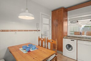 Una cocina compacta con paredes de azulejos blancos, una mesa de comedor de madera con tazas azules, una lavadora y una lámpara colgante.
