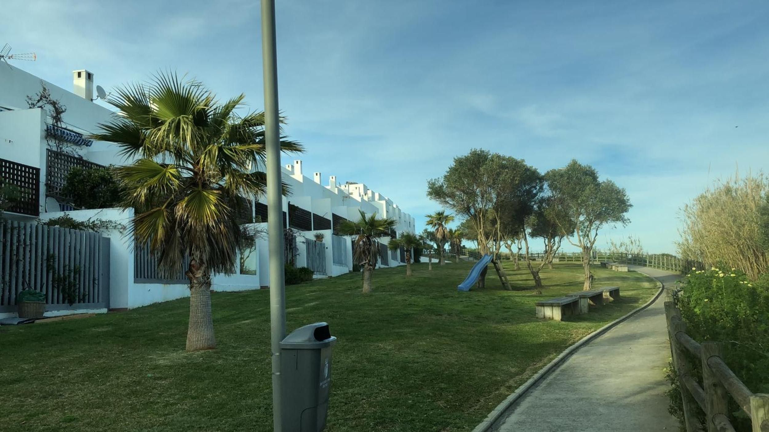 A pathway leading to a residential area with white buildings, palm trees, and a playground with a slide.