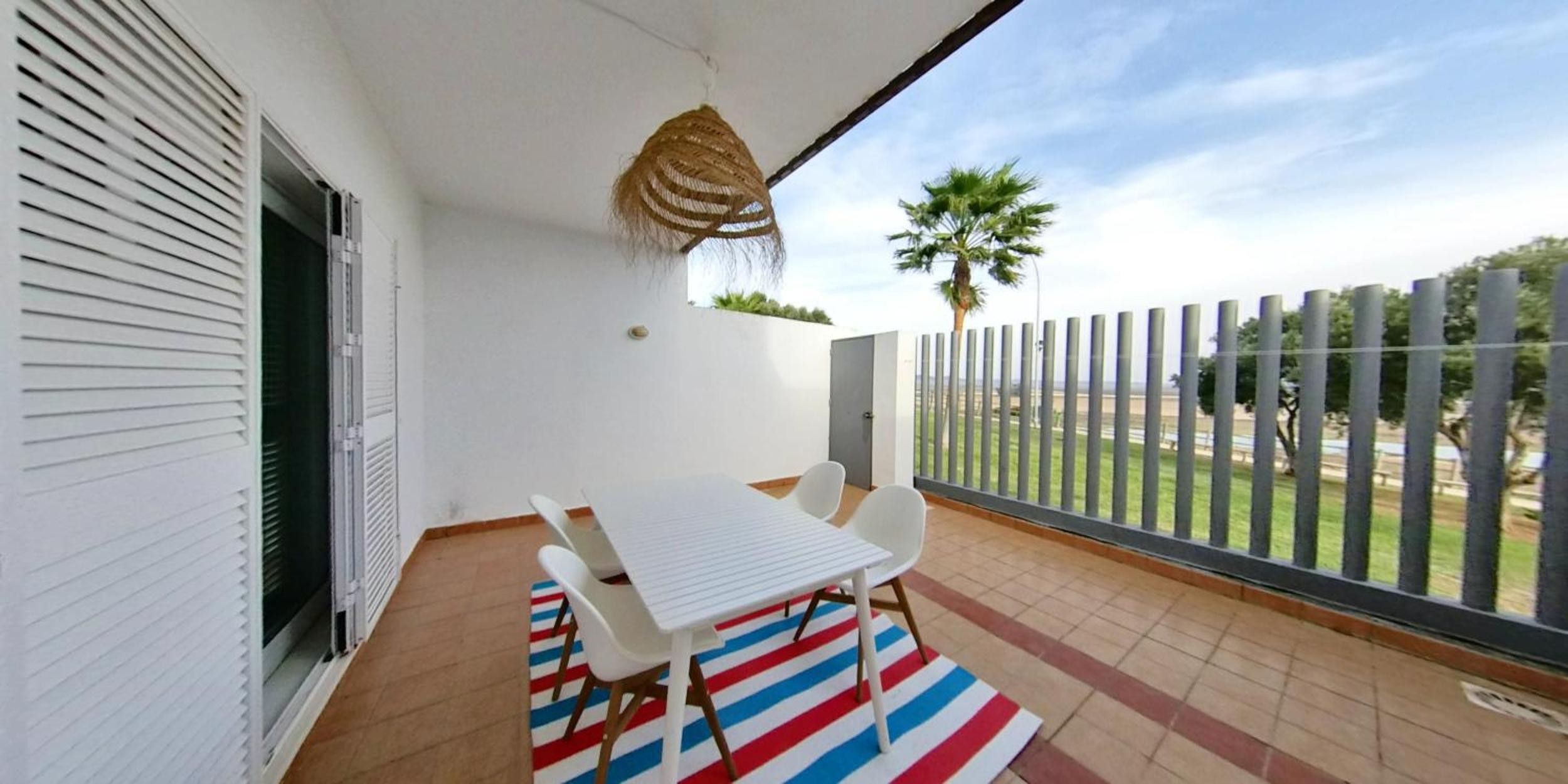 Un balcon avec une table et des chaises blanches, un tapis à rayures et une vue sur une clôture et de la verdure.