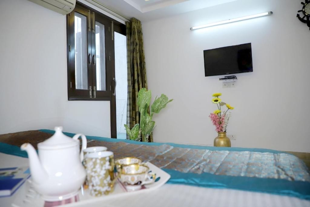 Une chambre d’hôtel avec un lit, un téléviseur à écran plat fixé au mur, une fenêtre avec des rideaux et une petite table sur laquelle se trouvent une théière et des tasses.