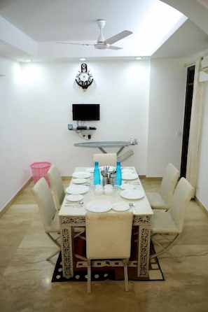 Uma sala de jantar com uma mesa posta para quatro pessoas, um ventilador de teto, uma TV fixada na parede e um aplique.