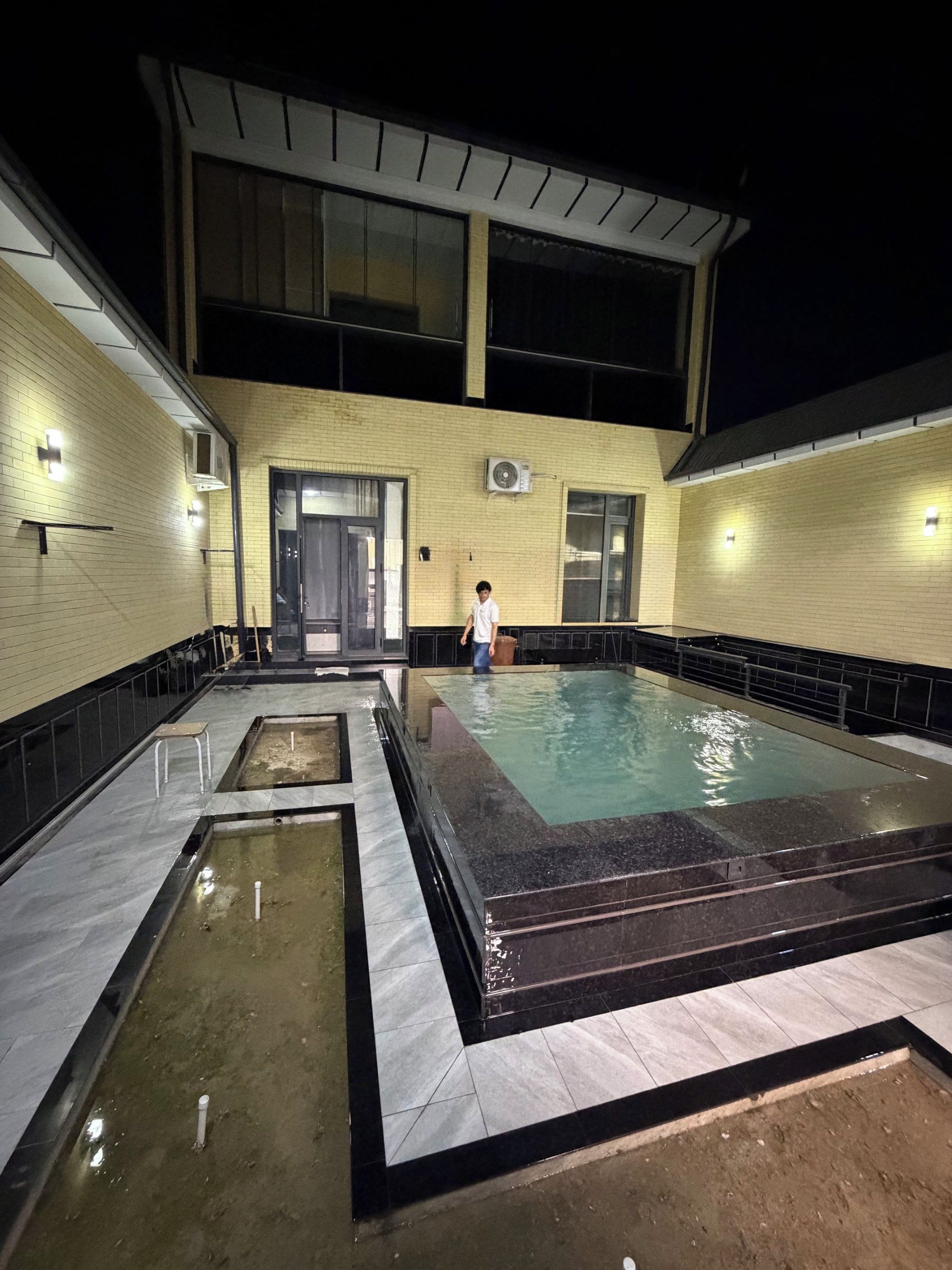 Área de piscina com deck revestido, uma pessoa próxima à piscina e um edifício com grandes janelas e unidade de ar condicionado.