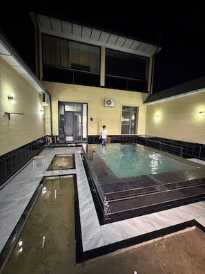 A swimming pool area with a tiled deck, a person standing by the pool, and a building with large windows and an air conditioning unit. - Happy Stay (Tashkent)