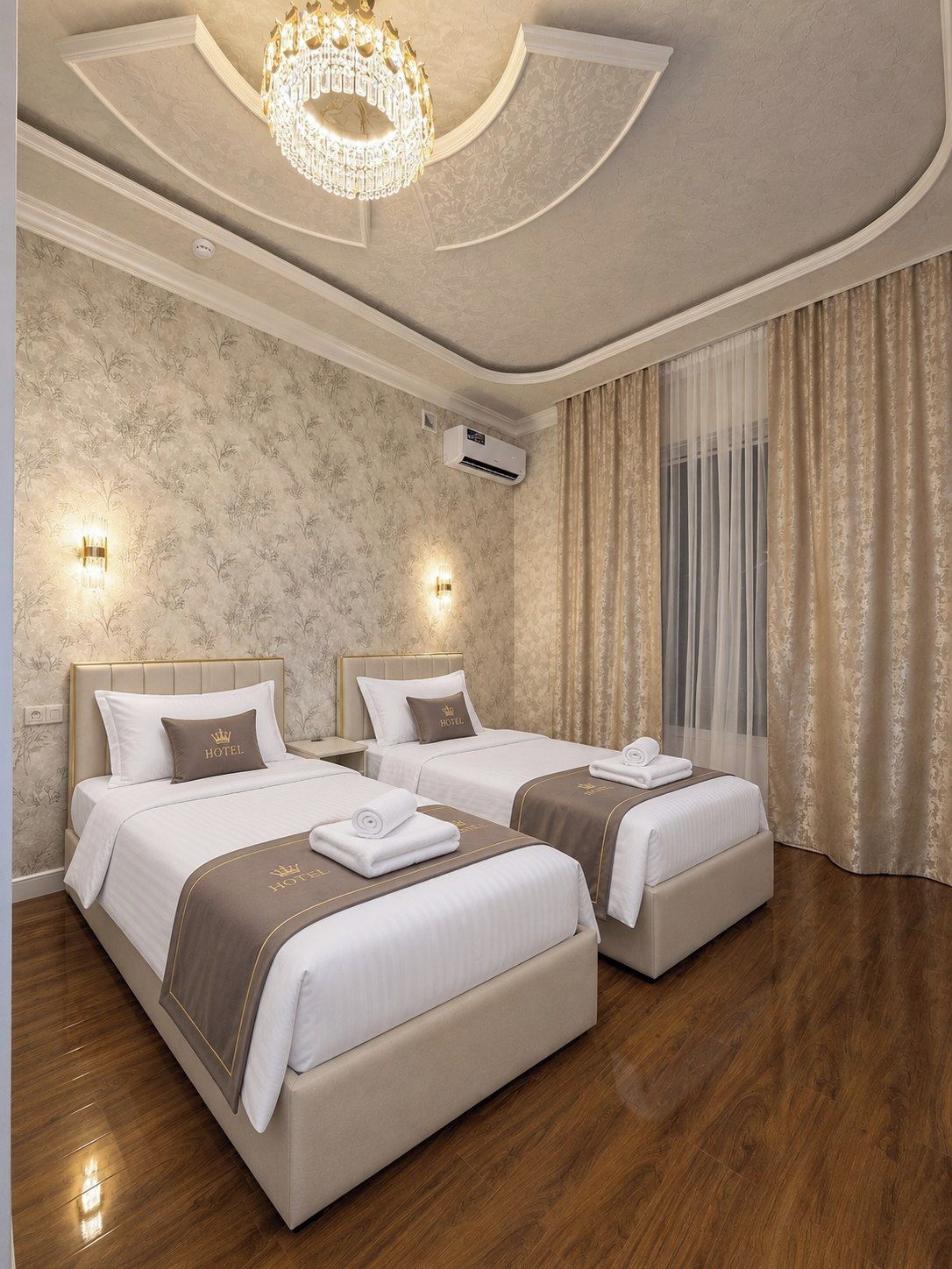 Quarto de hotel com duas camas, um lustre, arandelas e uma janela grande com cortinas.
