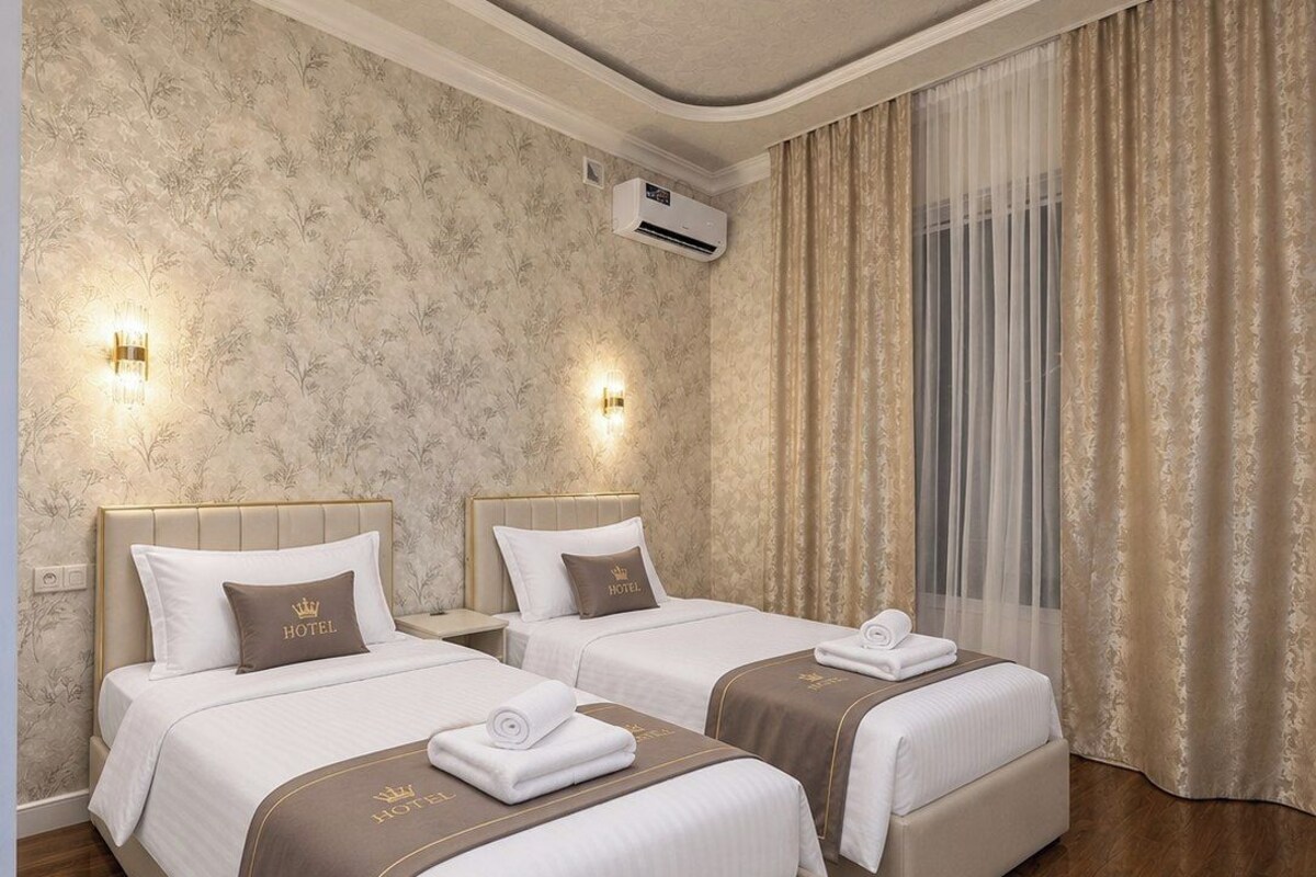 Quarto de hotel com duas camas, um lustre, arandelas e uma janela grande com cortinas.