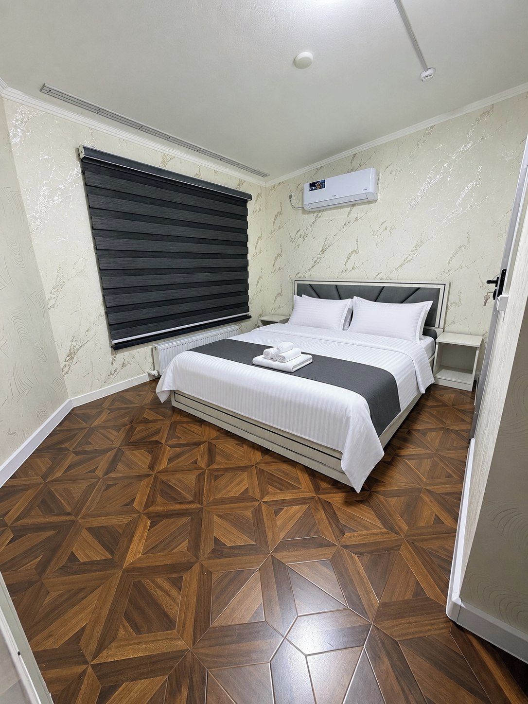 A bedroom with a bed, a window with blinds, an air conditioning unit, and a patterned wooden floor.
