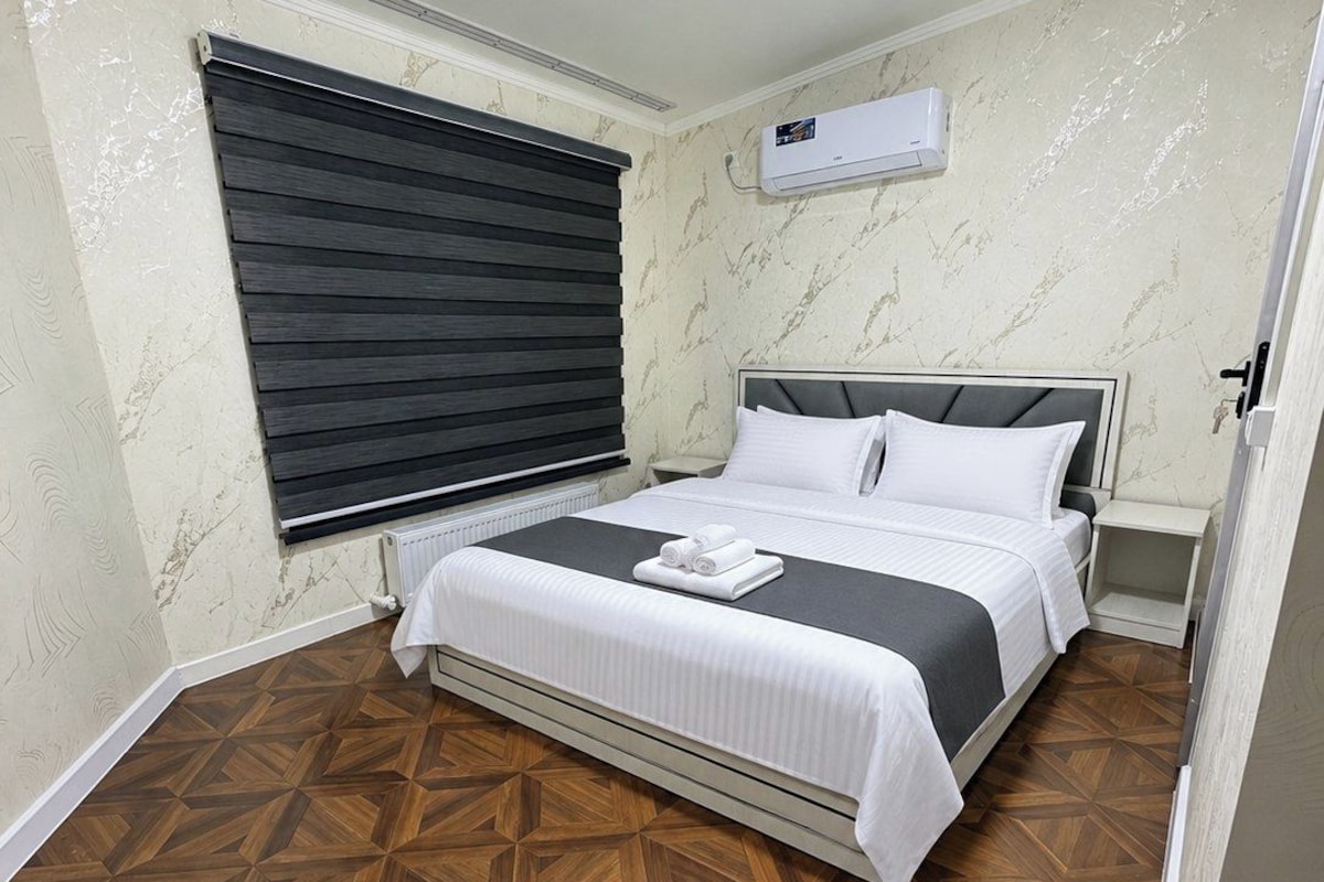 Quarto com cama, janela com persianas, ar condicionado e piso de madeira estampado.