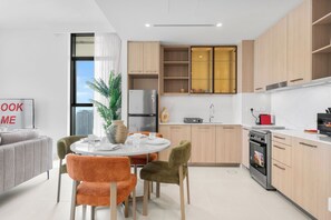 A modern kitchen with a dining area, featuring a refrigerator, oven, and a large window with a city view. - StayShort - Refined Classy 1 Bedroom in Dubai Hills - Sleeps 2! (Dubai Hills)