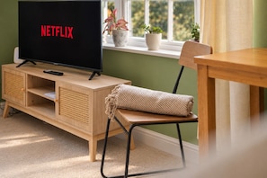 A room with a television displaying the Netflix logo, a wooden cabinet, a chair, a small table, and a window with plants. - Town centre l 1bd apt l 5 min to station (Hertfordshire)