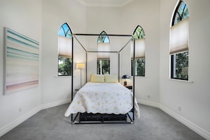 A four-poster bed with a canopy, a painting on the wall, and a view of trees through the windows. - Hemingway Place - Charming Naples Retreat (Naples)