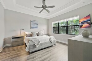 A bedroom with a bed, bedside tables, a ceiling fan, a window with a view of greenery, and a painting on the wall. - Esplanade Golf & Country Club (Naples)
