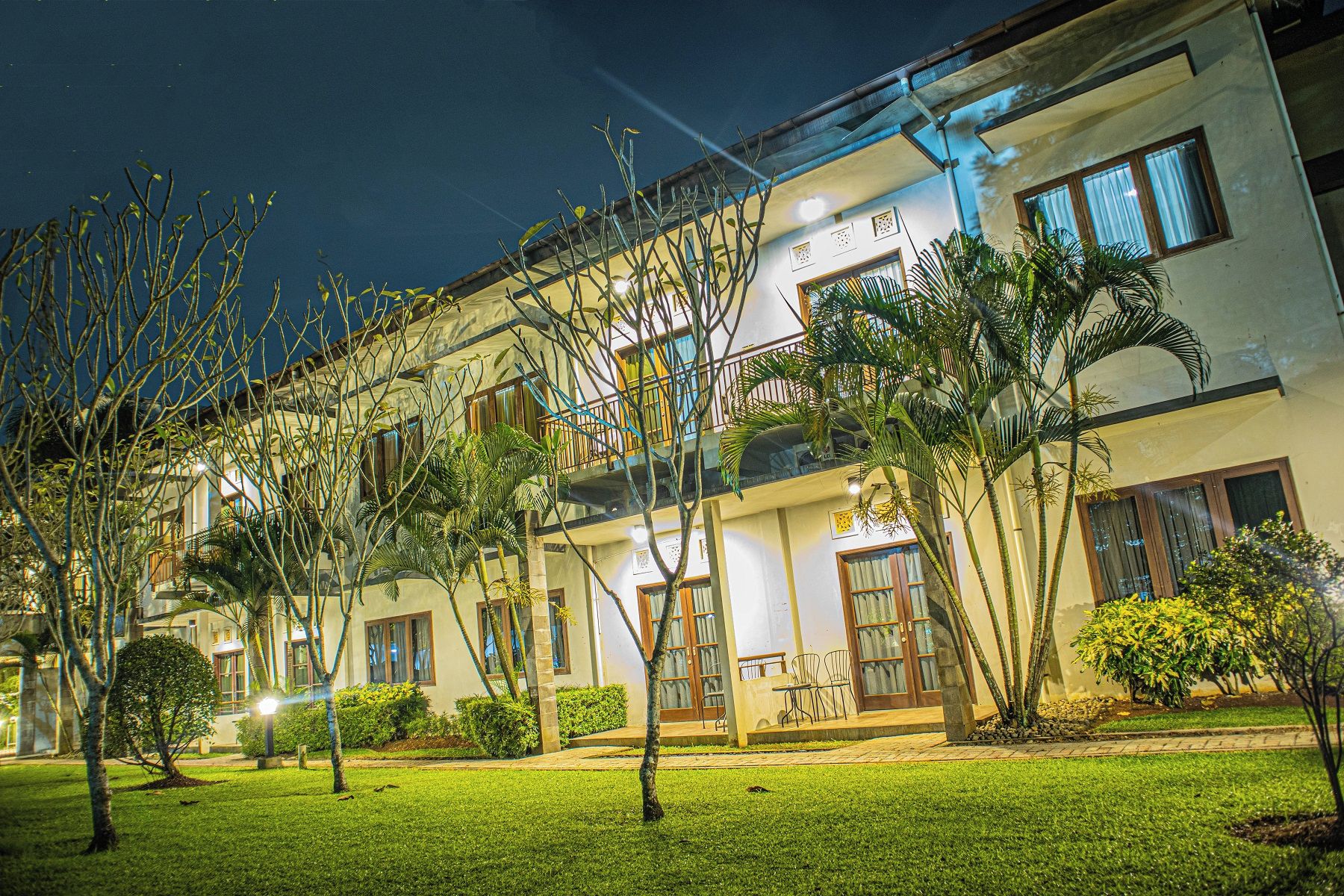 A multi-story hotel with a balcony on the second floor, surrounded by well-maintained lawns and various plants.