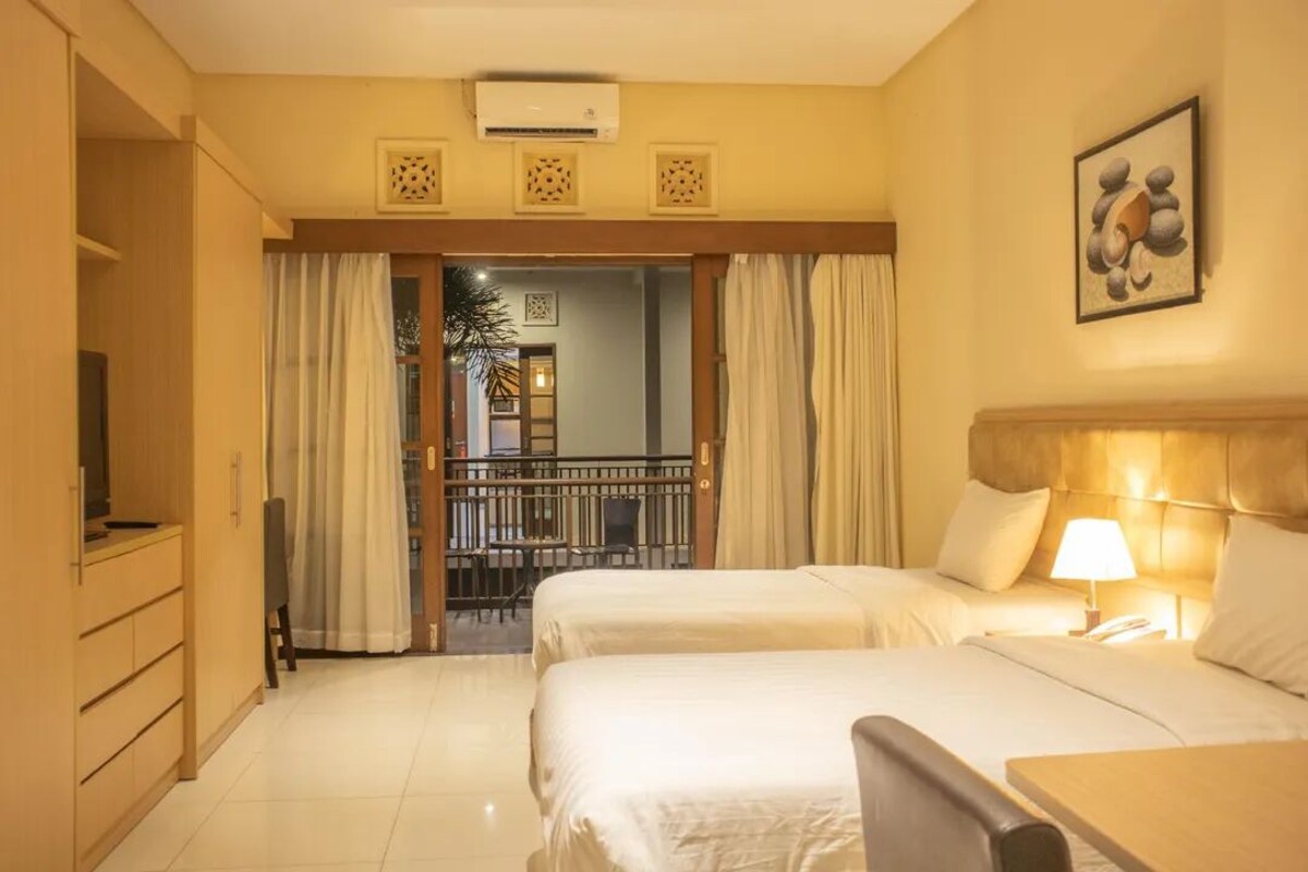 A hotel room with two beds, a desk, a chair, a TV, and a balcony with a railing.