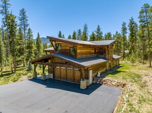 A two-story log cabin with a covered porch and a garage, surrounded by a forested area. - Tabernash Retreat | 3BR, Hot Tub, Mtn Views (Tabernash)