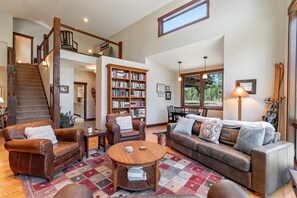 A spacious living room with a high ceiling, a staircase, a large bookshelf, leather furniture, and a view of the outdoors. - Tabernash Retreat | 3BR, Hot Tub, Mtn Views (Tabernash)