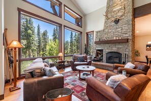 A spacious living room with a stone fireplace, leather furniture, and large windows offering a view of trees. - Tabernash Retreat | 3BR, Hot Tub, Mtn Views (Tabernash)