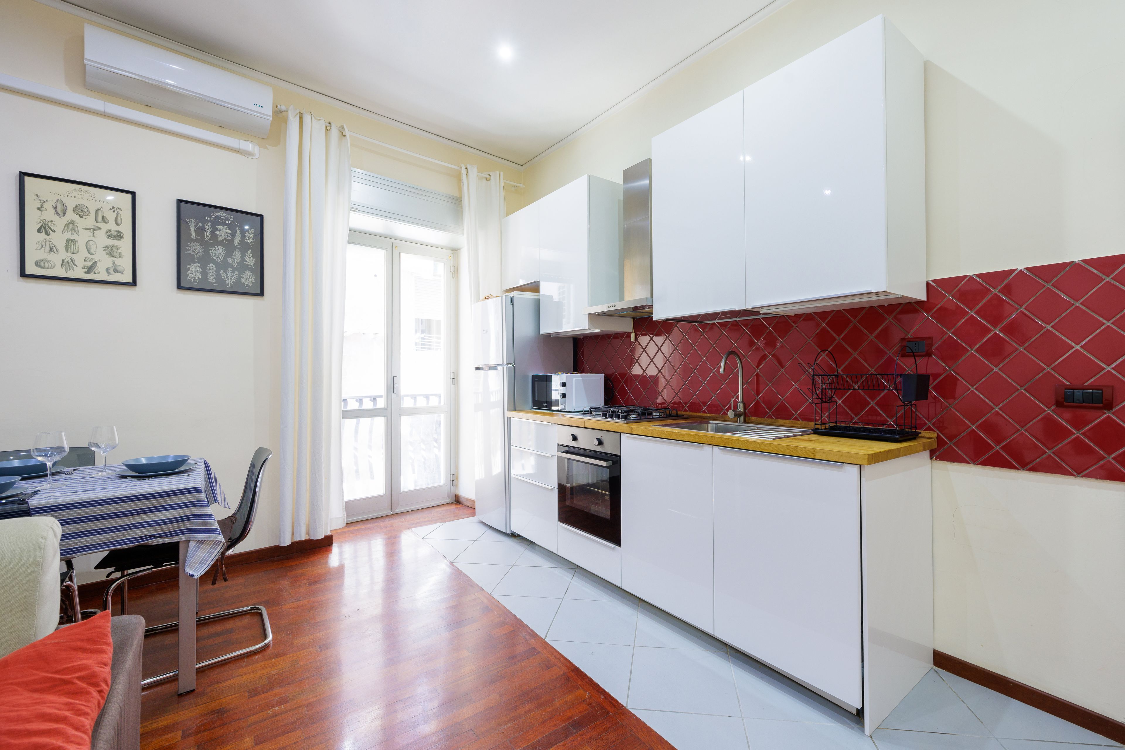 Une cuisine de taille réduite avec des meubles blancs, un pare-feu rouge et un coin repas agrémenté d’une nappe à rayures.