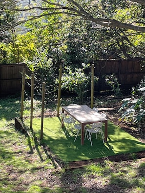 A wooden table with chairs in a garden area with green artificial turf, surrounded by plants and a wooden fence. - Charming cottage in the heart of Maleny. Two minute walk from the main street. (Maleny)