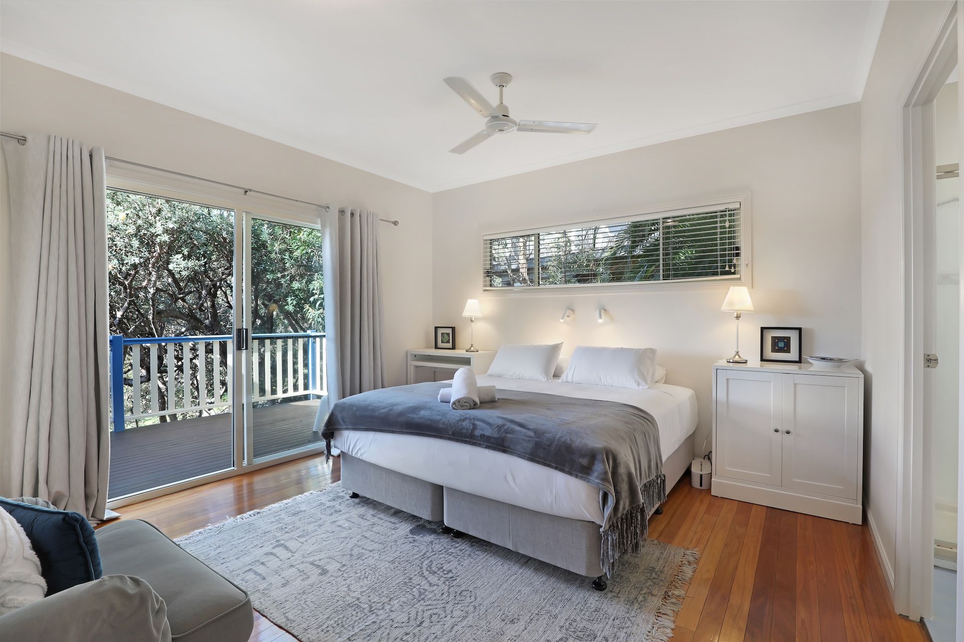 Ein Schlafzimmer mit Bett, Deckenventilator, Nachttisch, Lampe, einem Fenster mit Jalousien und einem Balkon mit Geländer.