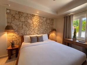 A bedroom with a stone wall, a bed with white linens, two bedside lamps, a nightstand, and a window with a view of greenery. - New 5-bedroom family villa in Esmeralda-noord, just 3 minutes from Arashi Beach. Enjoy peaceful Salinja views, a beautiful private pool, natural stone details, a covered outdoor patio with BBQ, 5 king bedrooms, a full kitchen,and space for 10 Guests (Noord)