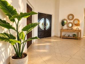 A hallway with a potted plant, a wooden console table with decorative items, and a double door with glass panels. - New 5-bedroom family villa in Esmeralda-noord, just 3 minutes from Arashi Beach. Enjoy peaceful Salinja views, a beautiful private pool, natural stone details, a covered outdoor patio with BBQ, 5 king bedrooms, a full kitchen,and space for 10 Guests (Noord)