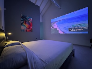 Ein Schlafzimmer mit einem Bett, einer Projektionsleinwand, die eine Strandansicht zeigt, und einem Gemälde an der Wand.