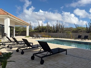 Uma área à beira da piscina com espreguiçadeiras, um espaço de estar coberto e vista para cactos e árvores.