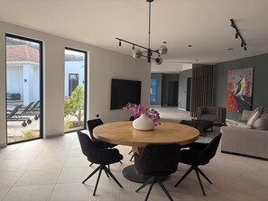 Uma área de jantar moderna com mesa redonda, cadeiras pretas, um vaso com flores e vista para uma piscina e área externa.