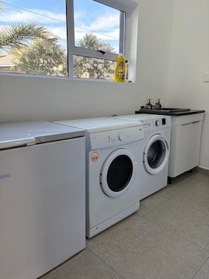 Une buanderie équipée d’un lave-linge et d’un sèche-linge, une fenêtre donnant sur des arbres et une bouteille de liquide vaisselle posée sur le rebord de la fenêtre.
