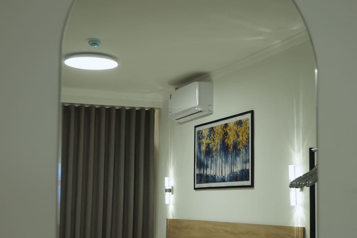 A hotel room with a bed, a headboard, a painting, an air conditioning unit, and a window with curtains.