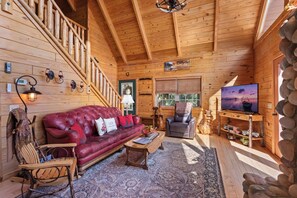 A log cabin interior with a red leather sofa, wooden staircase, and a television. - Mountain Views & Fire Pit | Dog-Friendly Cabin (Warne)