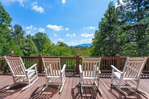 A wooden deck with white rocking chairs facing a forested landscape. - Mountain Views & Fire Pit | Dog-Friendly Cabin (Warne)