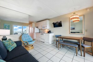 A modern living room with a sofa, a dining table with chairs, a kitchen area with appliances, and a large window offering a view. - Beacher's Oceanfront Balcony, Condo Complex Construction 10/2026-1/2027 (St. Augustine)