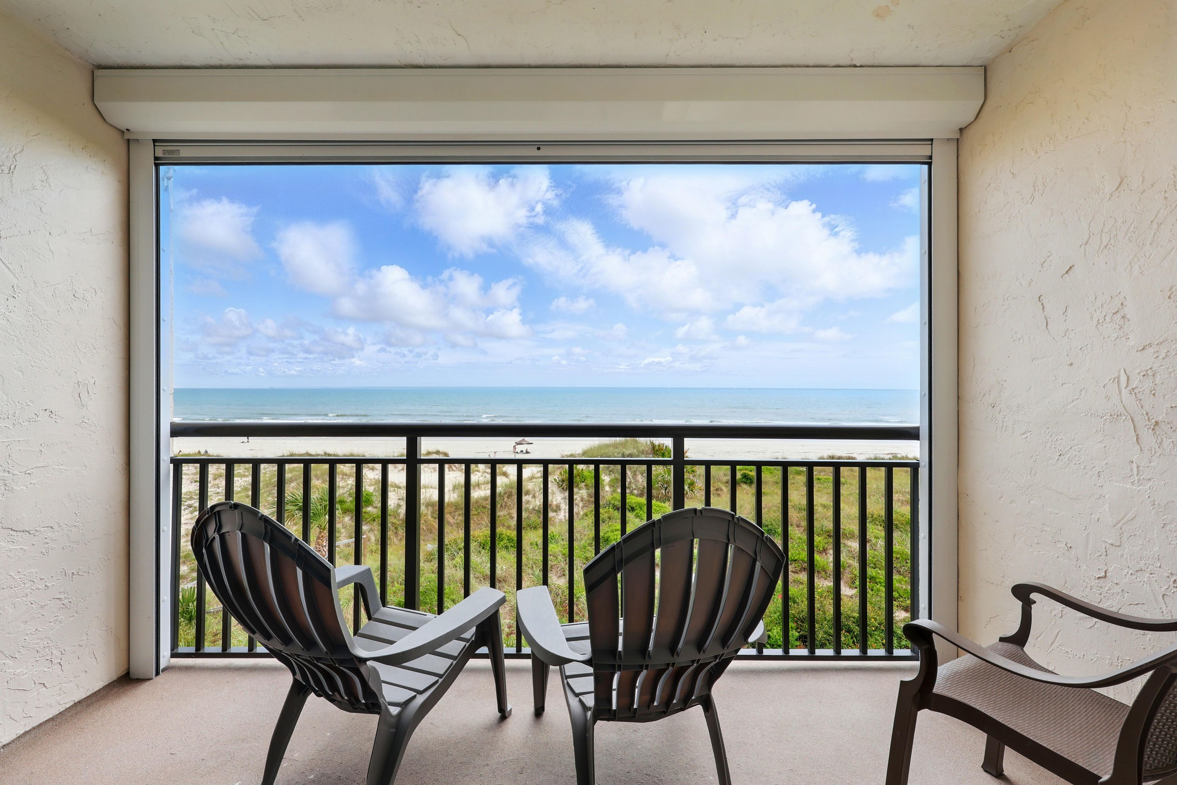 Un balcon avec deux chaises et une table, donnant sur une plage sous un ciel dégagé.