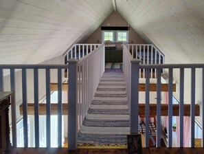 Eine Holztreppe mit weißen Geländern führt zu einem Schlafzimmer auf dem Dachboden, das ein Fenster hat.