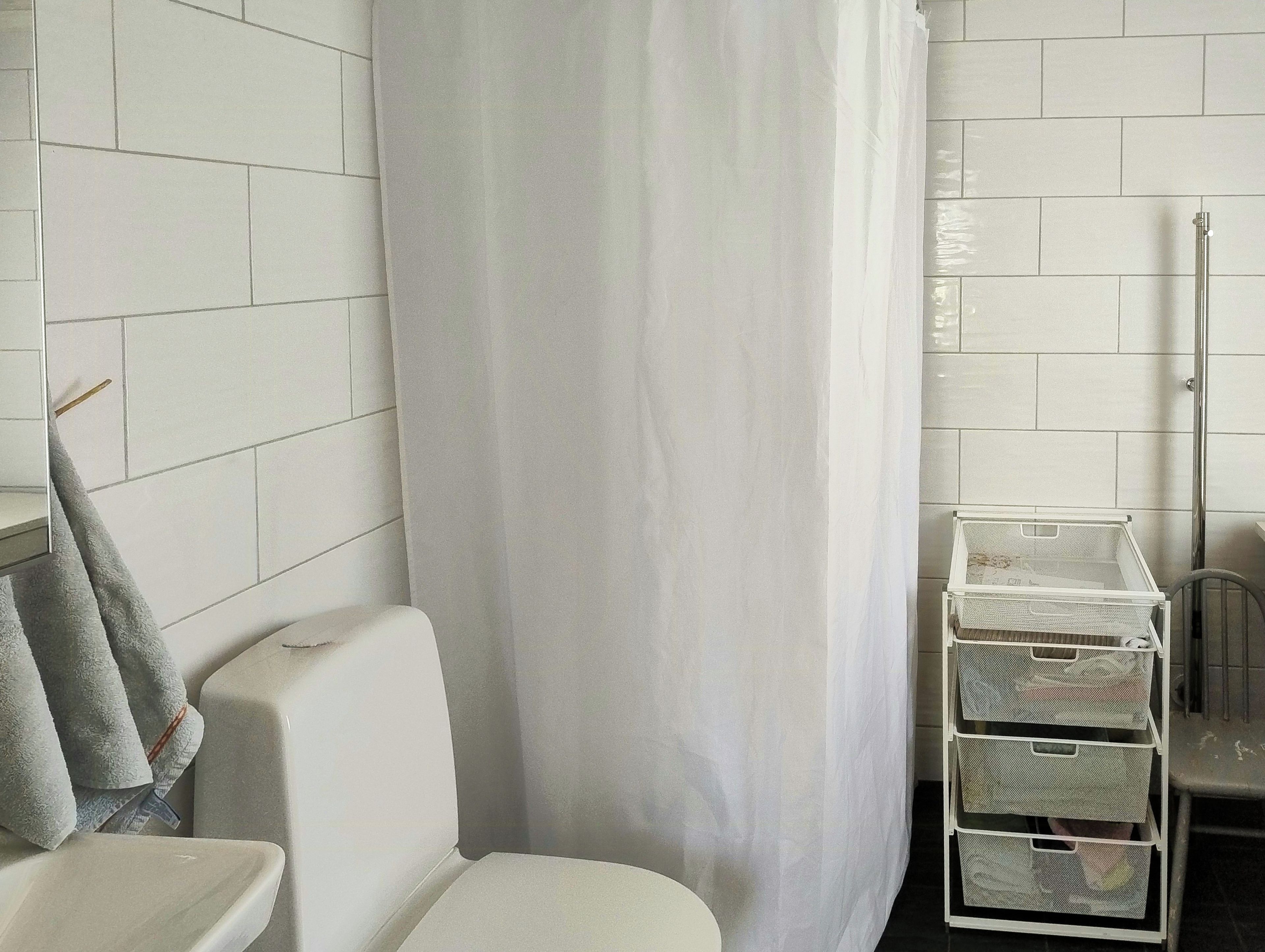 Une salle de bain avec des murs carrelés de carreaux blancs, des toilettes, un rideau de douche et un meuble de rangement en métal doté de tiroirs.