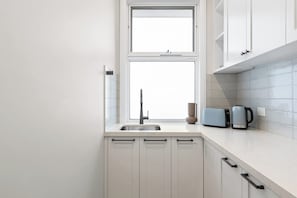 A kitchen with white cabinets, a double sink, a window, and a kettle on the counter. - Oakwood Retreat (Rosebud)