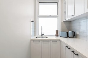 A kitchen with white cabinets, a double sink, a window, and a kettle on the counter. - Heated pool, Rosewood Oakwood  Retreat (Rosebud)