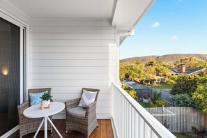 A balcony with a white railing, a wicker chair, a small round table, and a view of a residential area with trees and hills in the background. - Heated pool, Rosewood Oakwood  Retreat (Rosebud)