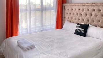 Quarto de hotel com uma cama grande, roupa de cama branca e cortinas laranja.