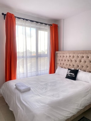 Quarto de hotel com uma cama grande, roupa de cama branca e cortinas laranja.