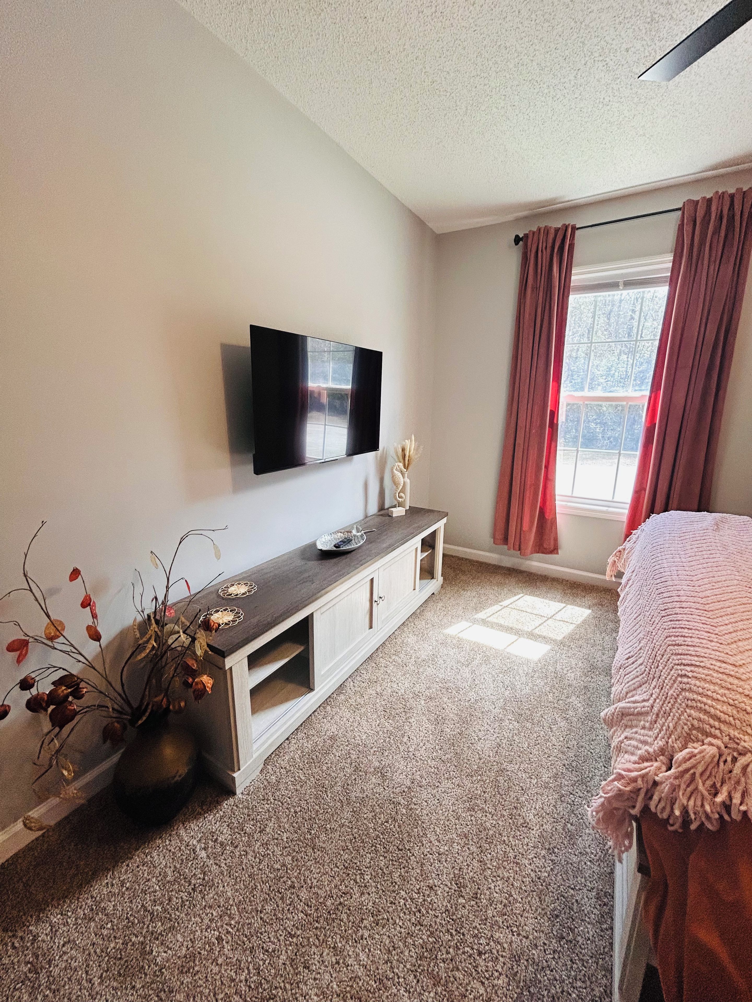 A bedroom with a bed, a TV mounted on the wall, a window with curtains, and a small plant on a console table.