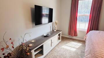 A bedroom with a bed, a TV mounted on the wall, a window with curtains, and a small plant on a console table.