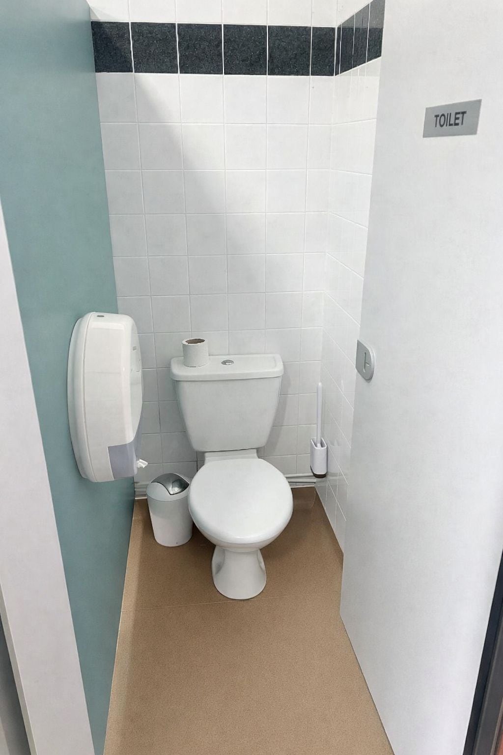 Un baño pequeño y limpio con inodoro, un cubo de basura y dispensador de jabón para manos.