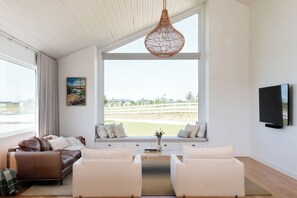 Ein geräumiges Wohnzimmer mit großem Fenster, einem braunen Ledersofa, einem weißen Sofa und einem an der Wand montierten Fernseher.